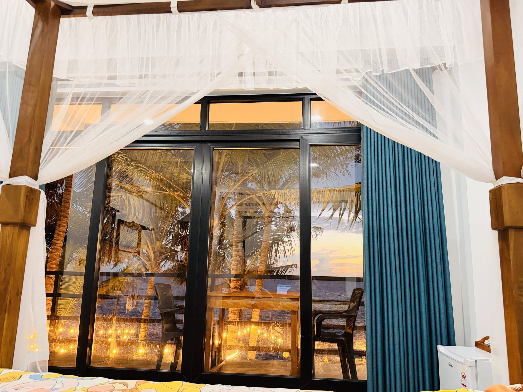A four-poster bed with white drapes, the glass doors open to palms and the sea at sunset